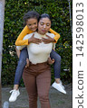 Mother carrying child on back smiling in backyard by swing chain wearing white sneakers 142598013