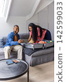 Couple packing open wheeled suitcase on gray sofa in loft, man holding tablet, passport visible 142599033