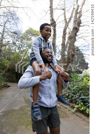 African American dad carrying son on shoulders walking paved park path in hoodies sneakers African American dad carrying son on shoulders walking paved park path in hoodies sneakers 142599410