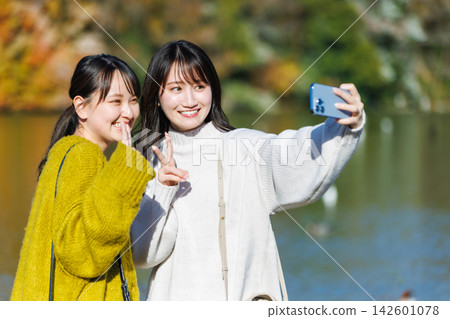 A young woman takes a selfie by the water's edge amidst the autumn foliage. 142601078