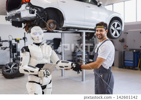 A smiling mechanic shakes hands with a humanoid robot holding a tablet in a modern auto repair shop with a car on a lift A smiling mechanic shakes hands with a humanoid robot holding a tablet in a modern auto repair shop with a car on a lift 142601142