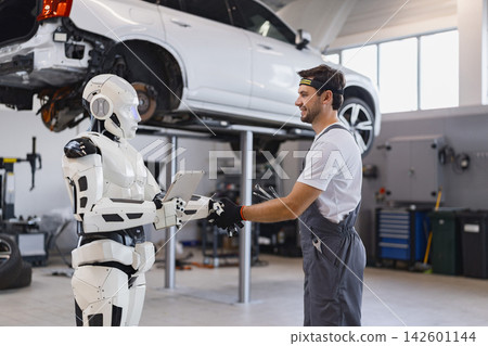 A human mechanic and a humanoid robot work together in an auto repair shop, discussing a vehicle on a lift A human mechanic and a humanoid robot work together in an auto repair shop, discussing a vehicle on a lift 142601144