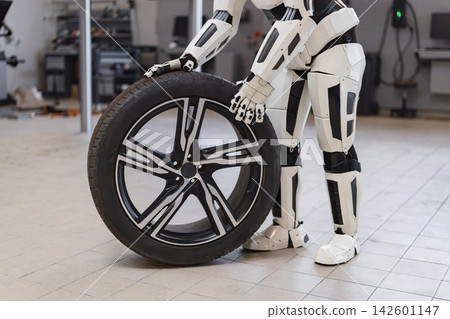 A humanoid robot in a workshop is holding a car tire and wheel, suggesting automated automotive repair A humanoid robot in a workshop is holding a car tire and wheel, suggesting automated automotive repair 142601147