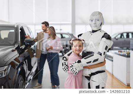 A young girl hugs a humanoid robot while a couple looks at a car in the background of a modern showroom A young girl hugs a humanoid robot while a couple looks at a car in the background of a modern showroom 142601210