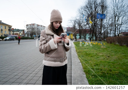 Young woman dressed warmly checks her phone in a city square during a chilly afternoon, surrounded by colorful flags and lush greenery Young woman dressed warmly checks her phone in a city square during a chilly afternoon, surrounded by colorful flags and lush greenery 142601257