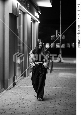 Woman strolling on a quiet city street at night, illuminated by soft urban lights, showcasing casual fashion with a relaxed attitude as she walks down the pavement Woman strolling on a quiet city street at night, illuminated by soft urban lights, showcasing casual fashion with a relaxed attitude as she walks down the pavement 142601261