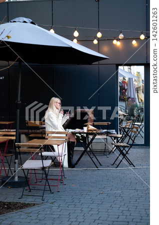 Charming outdoor cafe setting featuring a woman enjoying her time alone while sipping a beverage in a cozy ambiance on a breezy afternoon Charming outdoor cafe setting featuring a woman enjoying her time alone while sipping a beverage in a cozy ambiance on a breezy afternoon 142601263