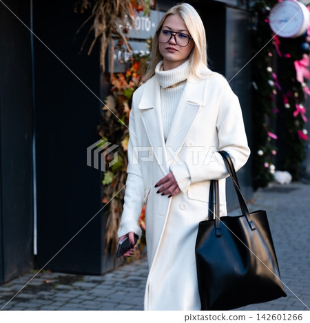 Chic woman in a stylish white coat strolling through a vibrant street adorned with autumn decor and unique shops on a crisp afternoon Chic woman in a stylish white coat strolling through a vibrant street adorned with autumn decor and unique shops on a crisp afternoon 142601266