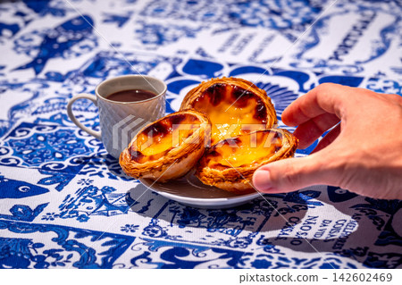 Hand picking Portuguese custard tart from white plate by small porcelain coffee cup with geometric pattern full of freshly-brewed espresso in full sunshine on blue and white tablecloth, with Hand picking Portuguese custard tart from white plate by small porcelain coffee cup with geometric pattern full of freshly-brewed espresso in full sunshine on blue and white tablecloth, with 142602469