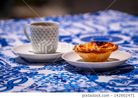 Close view of one pastel de nata custard tart on white plate and small porcelain coffee cup with geometric pattern, in hard sunlight on blue and white tablecloth with traditional Portuguese azulejo Close view of one pastel de nata custard tart on white plate and small porcelain coffee cup with geometric pattern, in hard sunlight on blue and white tablecloth with traditional Portuguese azulejo 142602471