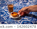 Hand adjusting a serving of single pastel de nata egg tart on white saucer beside glass of meia de leite coffee on blue and white tablecloth with azulejo motifs on sunny day in outdoor setting 142602478