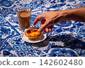 Hand reaching for a pastel de nata custard tart on white saucer beside glass of meia de leite coffee on blue and white tablecloth with azulejo motifs on sunny day, traditional Portuguese pastry, three 142602480