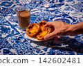 Three quarter view of hand picking famous dessert Portuguese custard tart from a serving on plate by glass of meia de leite coffee on blue and white tablecloth in warm outdoor light and blurred 142602481