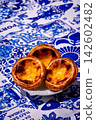 Three quarter shot of three pastel de nata custard tarts on white plate in hard sunlight on blue and white tablecloth with traditional Portuguese azulejo tile motifs. Glossy filling and golden flaky 142602482