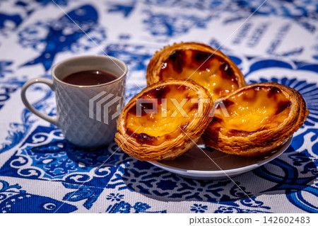 Three quarter shot of three pastel de nata custard tart on white plate and small porcelain coffee cup with geometric pattern, full of freshly-brewed espresso, outdoors on blue and white tablecloth Three quarter shot of three pastel de nata custard tart on white plate and small porcelain coffee cup with geometric pattern, full of freshly-brewed espresso, outdoors on blue and white tablecloth 142602483