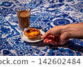 Hand placing a serving of single pastel de nata egg tart on white saucer beside glass of meia de leite coffee on blue and white tablecloth with azulejo motifs on sunny day, traditional Portuguese 142602484