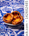 Three quarter shot of three pastel de nata custard tarts on white plate in hard sunlight on blue and white tablecloth with traditional Portuguese azulejo tile motifs. Crisp crust and juicy golden 142602485
