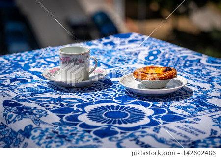 One pastel de nata custard tart on white plate and small porcelain coffee cup with floral pattern in bright sunlight on blue and white tablecloth with traditional Portuguese azulejo tile motifs 142602486