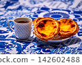 Three quarter shot of three pastel de nata custard tart on white plate and small porcelain coffee cup with geometric pattern, full of freshly-brewed espresso, in sunlight on blue and white tablecloth 142602488