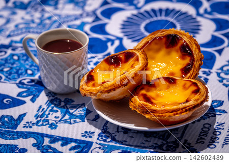 Three quarter shot of three pastel de nata custard tart on white plate and small porcelain coffee cup with geometric pattern, full of freshly-brewed espresso, on sunshine on blue and white tablecloth Three quarter shot of three pastel de nata custard tart on white plate and small porcelain coffee cup with geometric pattern, full of freshly-brewed espresso, on sunshine on blue and white tablecloth 142602489