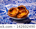 Three quarter shot of three pastel de nata custard tarts on white plate in hard sunlight on blue and white tablecloth with traditional Portuguese azulejo tile motifs. Glossy filling and golden flaky 142602490