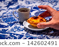 Hand grabbing Portuguese custard tart from white plate by small porcelain coffee cup, with geometric pattern full of freshly-brewed espresso in full sunlight, on blue and white tablecloth with 142602491