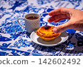 Hand reaching for Portuguese custard tart from white plate by small porcelain coffee cup with geometric pattern, full of freshly-brewed espresso, in total sunlight on blue and white tablecloth with 142602492