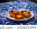 Three pastel de nata custard tarts on white plate on blue and white tablecloth with traditional Portuguese azulejo tile motifs. Iconic caramelized golden pastry in selective focus and high key light 142602499