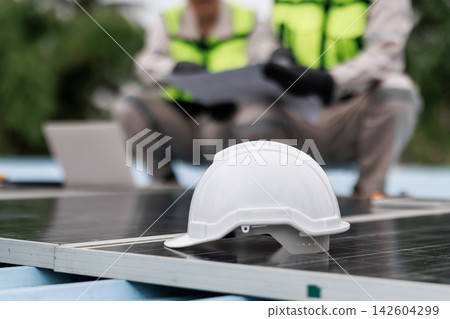White safety helmet resting on solar panels with two technicians in high visibility vests reviewing plans outdoors. White safety helmet resting on solar panels with two technicians in high visibility vests reviewing plans outdoors. 142604299