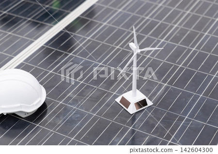 Solar panel surface with a white safety helmet and miniature wind turbine, clean energy, renewable power and sustainable construction. Solar panel surface with a white safety helmet and miniature wind turbine, clean energy, renewable power and sustainable construction. 142604300
