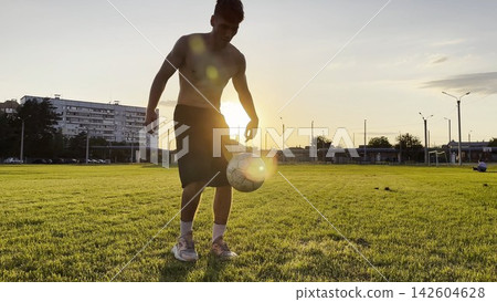 Young man juggling soccer ball on stadium at sunset. Professional footballer kicking ball at green field. Sportsman practicing tricks at meadow with sunlight at background. Freestyle football Young man juggling soccer ball on stadium at sunset. Professional footballer kicking ball at green field. Sportsman practicing tricks at meadow with sunlight at background. Freestyle football 142604628