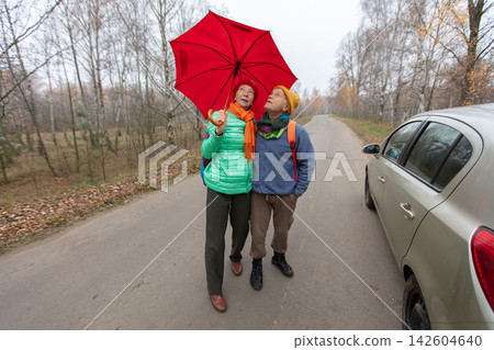 An elderly couple holds a red umbrella while standing on a road in the forest on a cloudy autumn day. 142604640