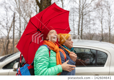 An elderly couple is walking together under a red umbrella on a cloudy day in a forest area wearing warm clothes. An elderly couple is walking together under a red umbrella on a cloudy day in a forest area wearing warm clothes. 142604641