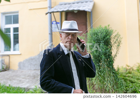 Man in formal attire wearing a hat speaks on a smartphone outside a yellow building during the day Man in formal attire wearing a hat speaks on a smartphone outside a yellow building during the day 142604696