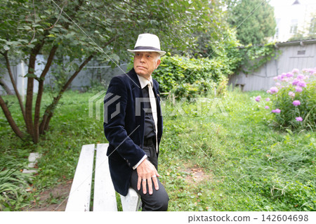 Man in elegant attire sitting on a bench in a lush garden surrounded by greenery and colorful flowers during daylight Man in elegant attire sitting on a bench in a lush garden surrounded by greenery and colorful flowers during daylight 142604698