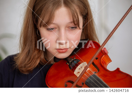 Young girl plays violin in a bright room during practice session in afternoon Young girl plays violin in a bright room during practice session in afternoon 142604716