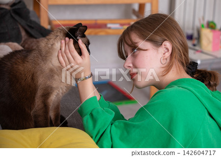 A girl sits on a couch and gently plays with a cat in a living room filled with stuffed toys and warm lighting during the afternoon. A girl sits on a couch and gently plays with a cat in a living room filled with stuffed toys and warm lighting during the afternoon. 142604717