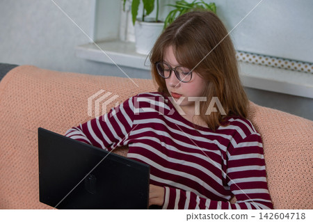 Girl using laptop while sitting on sofa in a cozy room with plants, focusing on screen during afternoon Girl using laptop while sitting on sofa in a cozy room with plants, focusing on screen during afternoon 142604718