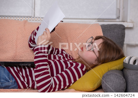 Girl reading notes while lying on a couch with a laptop nearby in a cozy indoor space during the afternoon Girl reading notes while lying on a couch with a laptop nearby in a cozy indoor space during the afternoon 142604724