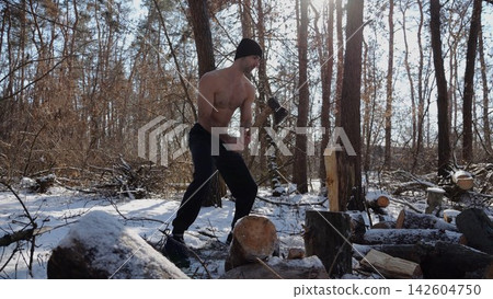 Shirtless young man chopping firewood in a sunny winter forest. Muscular guy working with axe in snowy woodland. Concept of strength, masculine, energy and harmony with nature during cold season 142604750