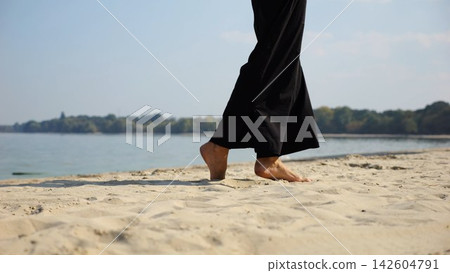Legs of adult woman stepping along sea shore. Female feet walking on sand at the beach with seascape at background. Barefoot lady strolling near ocean coast. Concept of summer vacation Legs of adult woman stepping along sea shore. Female feet walking on sand at the beach with seascape at background. Barefoot lady strolling near ocean coast. Concept of summer vacation 142604791