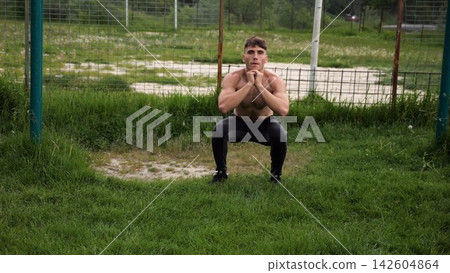 Male shirtless athlete doing squats at sports ground. Young sportsman working at summer nature. Strong sporty man exercising outdoor. Concept of active and sportive lifestyle. Slow motion Male shirtless athlete doing squats at sports ground. Young sportsman working at summer nature. Strong sporty man exercising outdoor. Concept of active and sportive lifestyle. Slow motion 142604864