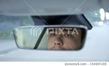 Male eyes of driver reflecting in rearview mirror while riding modern electric car. Young man looking concentrated to urban road during operating motor vehicle capturing focused gaze during city trip Male eyes of driver reflecting in rearview mirror while riding modern electric car. Young man looking concentrated to urban road during operating motor vehicle capturing focused gaze during city trip 142605108