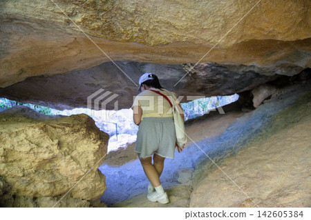 Woman hiker in a cap crouching under a low rock overhang on a canyon trail Woman hiker in a cap crouching under a low rock overhang on a canyon trail 142605384