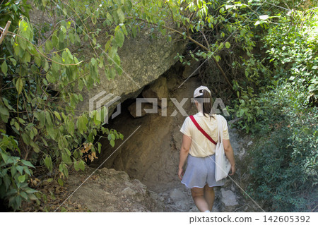 Woman hiker walking towards a cave entrance under a boulder on a woodland trail Woman hiker walking towards a cave entrance under a boulder on a woodland trail 142605392