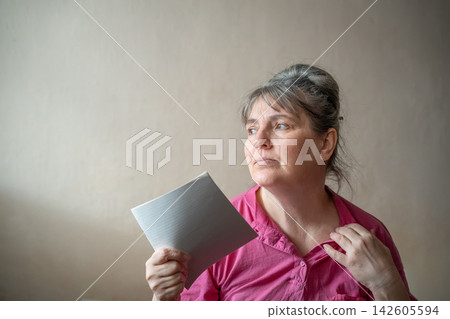 Mature woman cooling herself with a handheld paper fan at home. Menopause awareness hot flashes women's health and perimenopause wellbeing Mature woman cooling herself with a handheld paper fan at home. Menopause awareness hot flashes women's health and perimenopause wellbeing 142605594