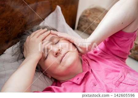 Mature woman resting in bed with hands on her forehead at home. Menopause awareness headache fatigue discomfort and recovery Mature woman resting in bed with hands on her forehead at home. Menopause awareness headache fatigue discomfort and recovery 142605596
