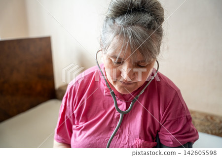Mature woman in pink top checking her wellbeing with a stethoscope at home. Menopause health awareness selfcare and climacteric support Mature woman in pink top checking her wellbeing with a stethoscope at home. Menopause health awareness selfcare and climacteric support 142605598
