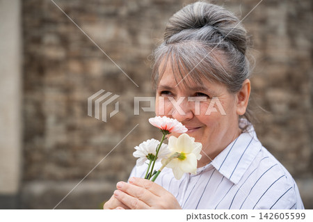 Mature woman smelling fresh flowers outdoors beside a stone wall. Menopause wellness positive aging aromatherapy and natural selfcare Mature woman smelling fresh flowers outdoors beside a stone wall. Menopause wellness positive aging aromatherapy and natural selfcare 142605599