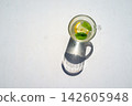 Top view of clear citrus drink in glass mug with lemon slice and mint leaves, casting long shadow on bright white surface, minimalist refreshment with clean summer feeling 142605948